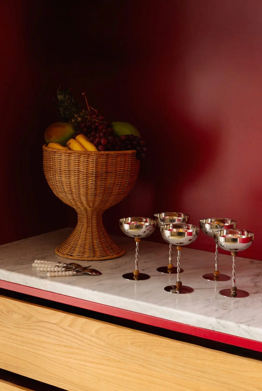 Beth Decoration Bowl styled on marble shelf with fruit, alongside silver coupe glasses on dark red background